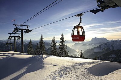 Eine Seilbahn fährt über eine schneebedeckte Landschaft. Im Hintergrund sind majestätische Berge und ein klarer Himmel zu sehen.