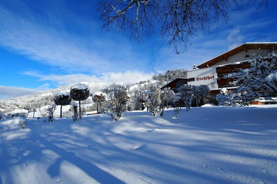 Verschneite Außenanlage des Hotels Bichlhof mit Bäumen und Bergen im Hintergrund unter blauem Himmel.