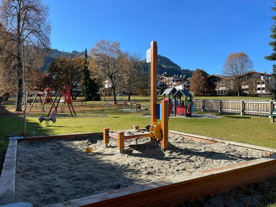 Spielplatz Pfarrau Ein Sandkasten mit Holzumrandung und Spielgeräten auf einer Wiese vor Bergen.
