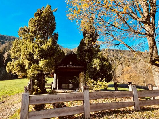 Kapelle am Kaiserblickweg Kleine Holzkapelle hinter einem Holzzaun, umgeben von Nadelbäumen und herbstlichem Laub vor bewaldetem Berg.