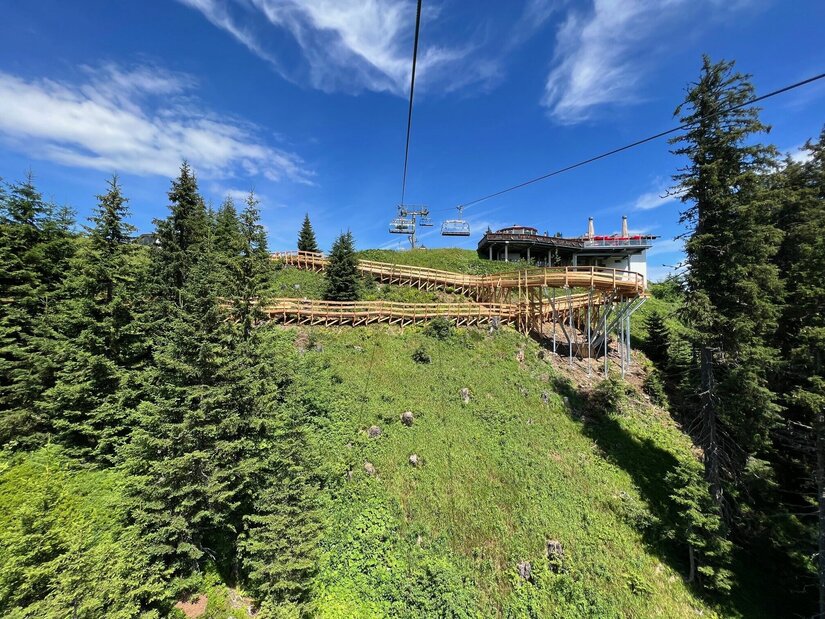 Upper Streiteck Trail Infrastruktur Blick auf einen hölzernen Steg am Berghang mit Sessellift und Nadelbäumen unter blauem Himmel.
