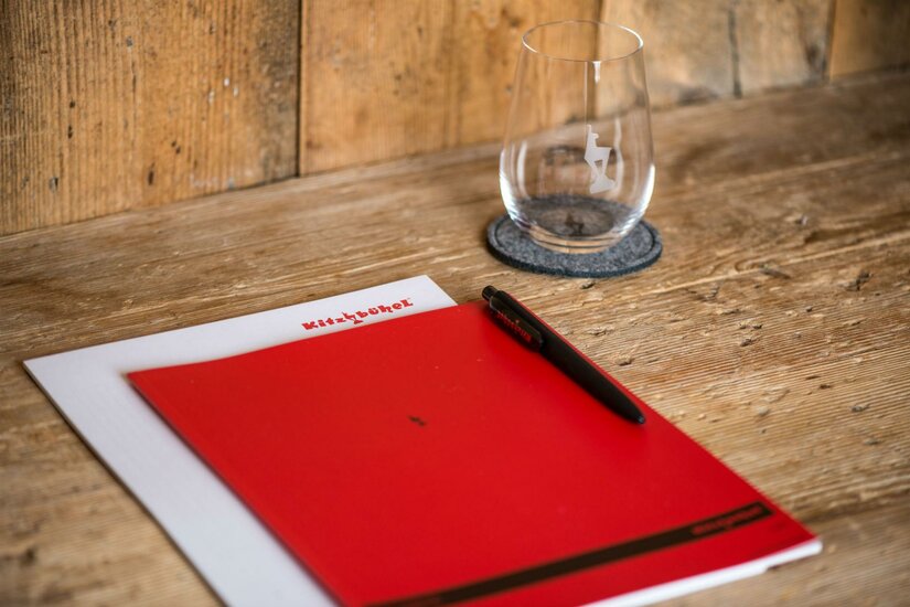 Ein rotes Notizbuch und ein Stift liegen auf einem Holztisch. Neben ihnen steht ein Glas auf einem Untersetzer.
