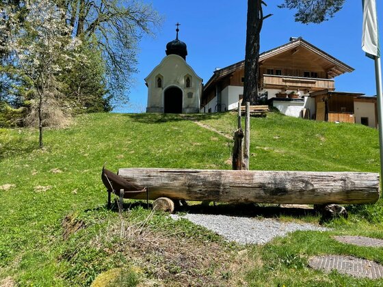 Kapelle und Holzbrunnen Holzbrunnen auf einer Wiese vor einer kleinen Kapelle und einem Holzhaus unter blauem Himmel.