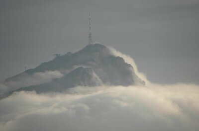Ein Berggipfel mit Sendemast ragt aus einer dichten Wolkendecke unter grauem Himmel hervor.