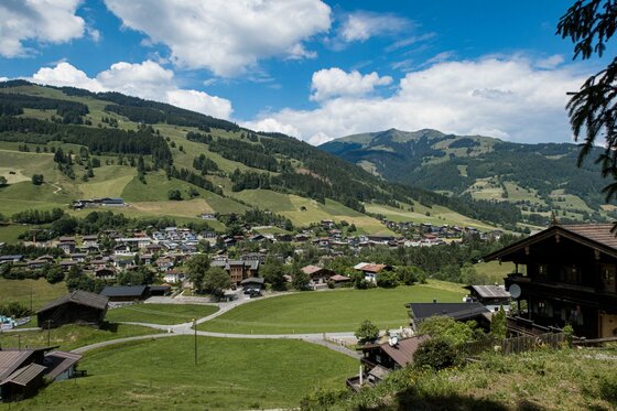 Dorfansicht Perlberg Tour Blick auf ein Dorf im Tal mit umliegenden bewaldeten Bergen und grünen Wiesen unter blauem Himmel.