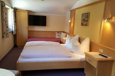 Dreibettzimmer im Gästehaus Wallner Helles Gästezimmer mit Doppelbett, Einzelbett im Hintergrund, Holzmöbeln und Flachbildfernseher an der Wand.