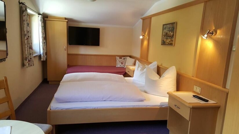 Dreibettzimmer im Gästehaus Wallner Helles Gästezimmer mit Doppelbett, Einzelbett im Hintergrund, Holzmöbeln und Flachbildfernseher an der Wand.