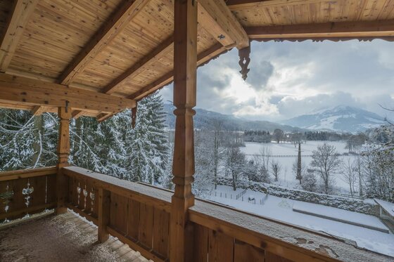 Blick von einem hölzernen Balkon auf eine verschneite Tallandschaft mit Bergen unter bewölktem Himmel.