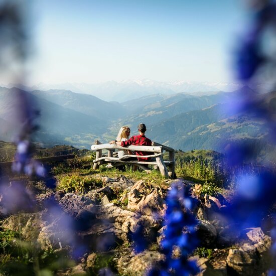 Ein Paar sitzt auf einer Holzbank und genießt die atemberaubende Aussicht auf die Berge und das Tal im Hintergrund.