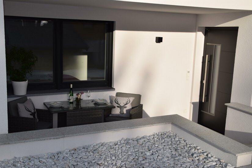 Moderne Terrasse mit Sitzgruppe, gedecktem Tisch und Kissen mit Hirschmotiv vor einer weißen Hauswand.