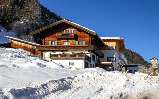 Alpiner Ferienhof mit Holzfassade und Balkonen in einer verschneiten Winterlandschaft unter blauem Himmel.