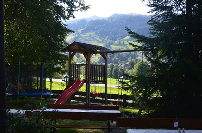 Holzspielturm mit roter Rutsche und Schaukel vor bewaldeter Bergkulisse.