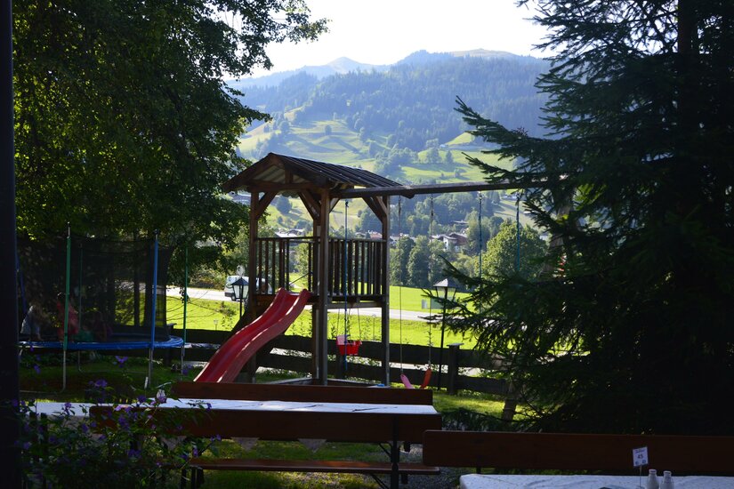 Holzspielturm mit roter Rutsche und Schaukel vor bewaldeter Bergkulisse.