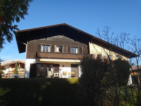 Zweistöckiges Gebäude mit dunkler Holzfassade, Balkon und Terrasse vor blauem Himmel.