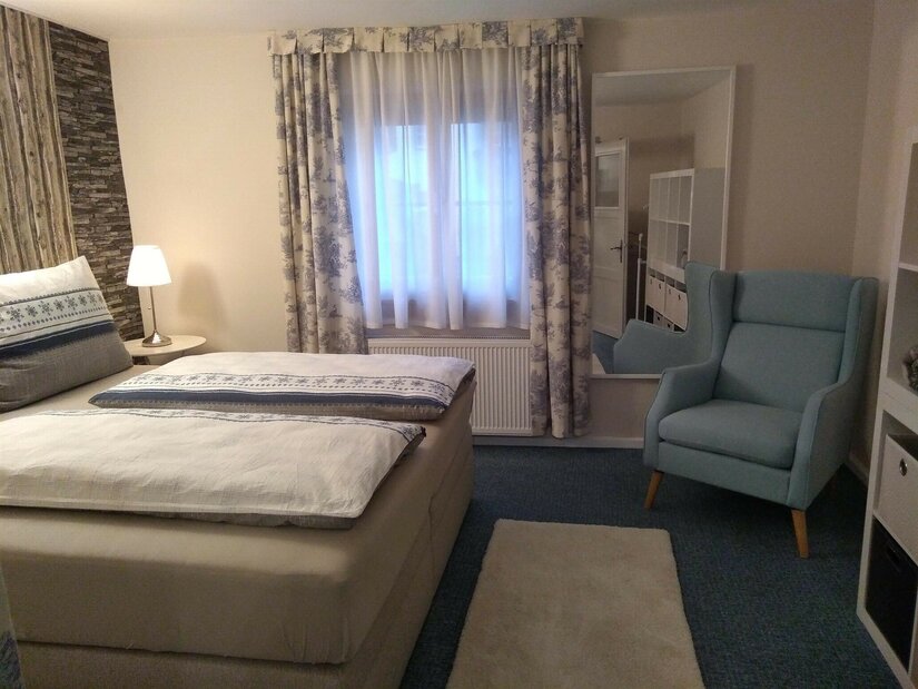 Kitzbüheler Suite Schlafzimmer Schlafzimmer mit Doppelbett, blauem Ohrensessel, Fenster mit Vorhängen und einem großen Spiegel.