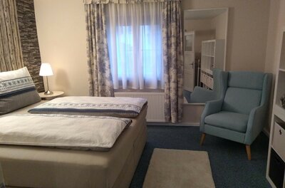 Schlafzimmer mit Doppelbett, blauem Ohrensessel, Fenster mit Vorhängen und einem großen Spiegel.