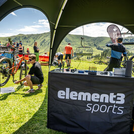 Ein Mechaniker arbeitet an einem Fahrrad unter einem Zelt bei einem Bike-Event in Kitzbühel, während im Hintergrund Menschen auf einer Wiese entspannen.