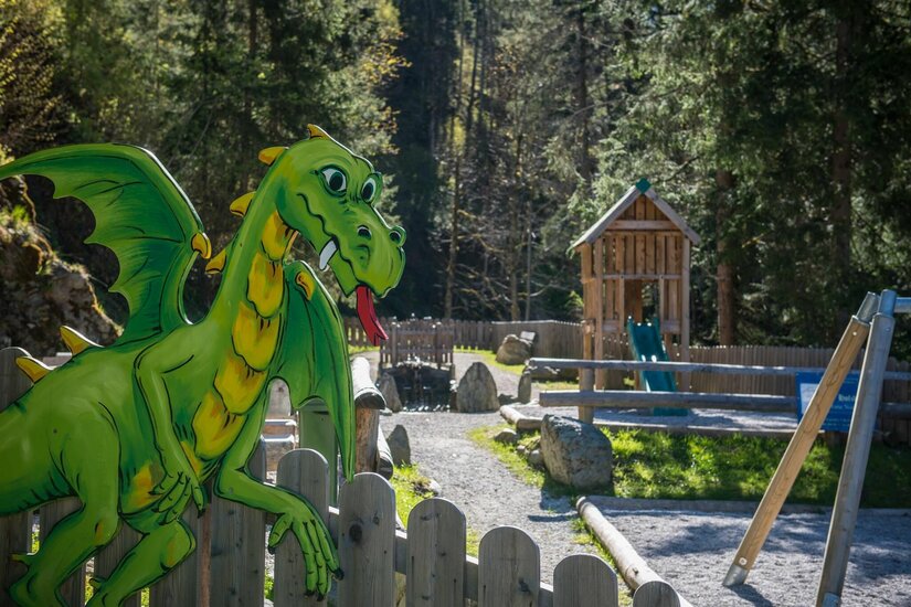 Grüne Drachenfigur aus Holz an einem Zaun vor einem Waldspielplatz mit Rutsche.