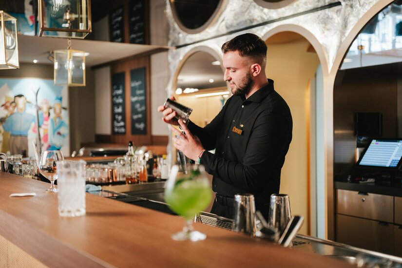 Grand Tirolia Cooper Bar Ein Barkeeper in schwarzer Kleidung bereitet hinter einem Holztresen einen Cocktail mit einem Shaker zu.