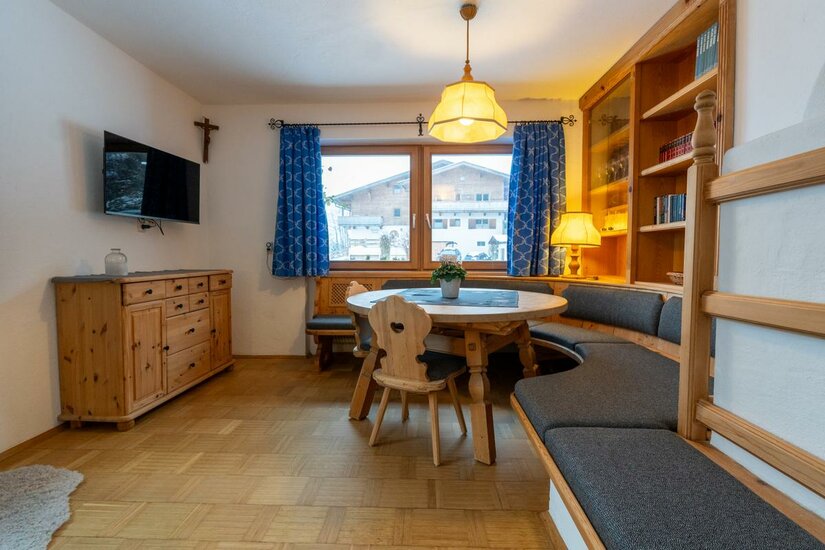 paulo korrektur 3-2 Ein gemütlicher Wohnraum mit einer Sitzecke aus Holz und einem runden Tisch. Große Fenster mit blauen Vorhängen bieten einen Blick auf die Außenansicht.