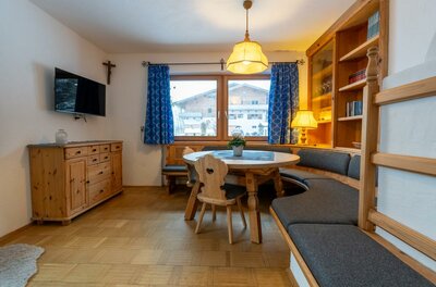 Ein gemütlicher Wohnraum mit einer Sitzecke aus Holz und einem runden Tisch. Große Fenster mit blauen Vorhängen bieten einen Blick auf die Außenansicht.