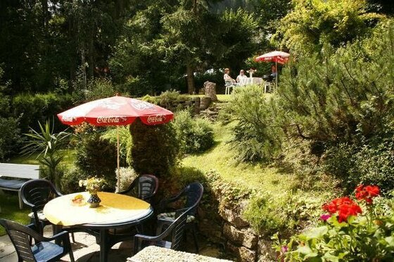 Garten der Pension Schatz Gartenterrasse mit runden Tischen, roten Sonnenschirmen und grüner Bepflanzung.