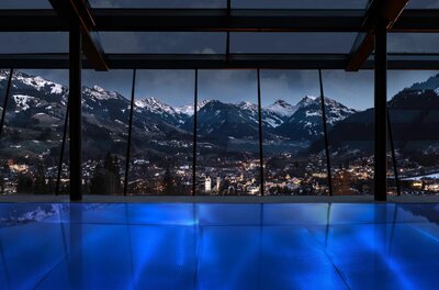 Pool im Lebenberg Schlosshotel Blick aus einem beleuchteten Innenpool durch eine Glasfront auf ein nächtliches Bergpanorama und eine beleuchtete Stadt.
