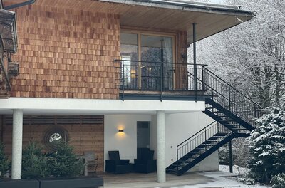 Ein modernes Haus mit Holzverkleidung und großen Fenstern. Der Garten ist schneeüberdeckt und im Hintergrund sind schneebedeckte Bäume sichtbar.