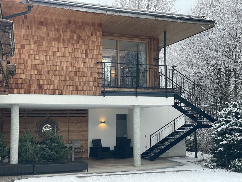 Ein modernes Haus mit Holzverkleidung und großen Fenstern. Der Garten ist schneeüberdeckt und im Hintergrund sind schneebedeckte Bäume sichtbar.