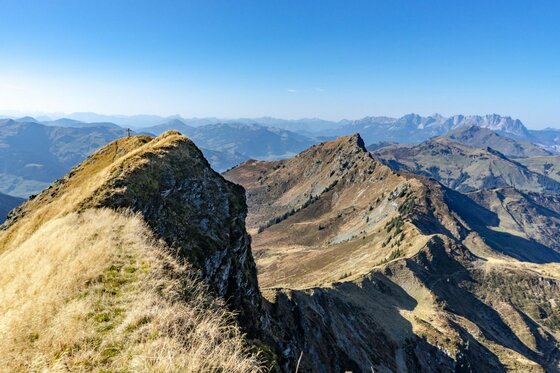 Blick auf einen schmalen Graskamm und felsige Berggipfel unter blauem Himmel.