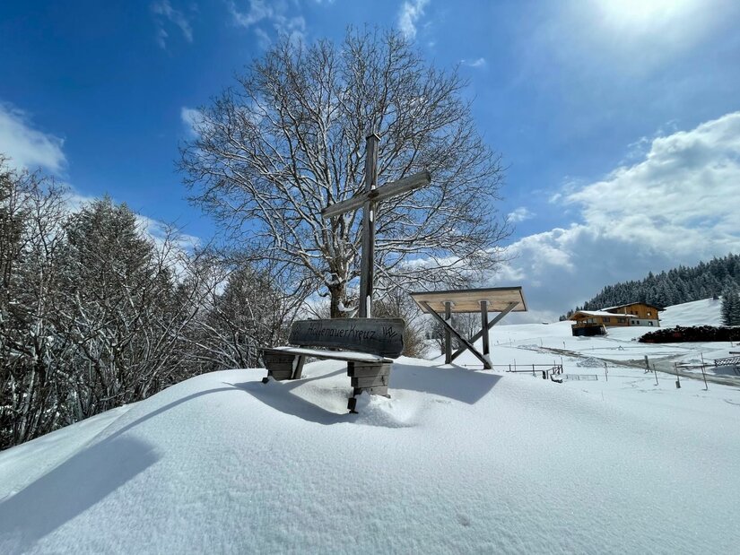 Holzkreuz mit Sitzbank auf einem verschneiten Hügel vor einem Baum und blauem Himmel.