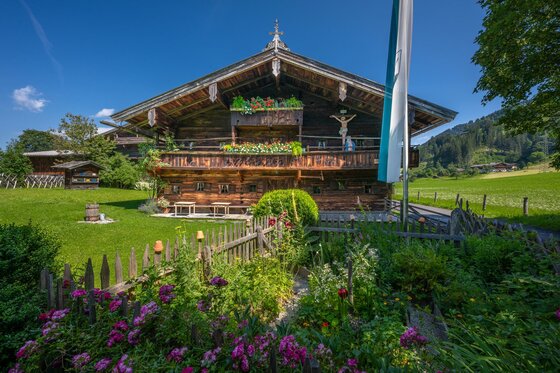 Ein traditionelles alpenländisches Haus mit Holzbalkon und bunten Blumen im Vordergrund. Die Umgebung ist grün und es herrscht ein klarer blauer Himmel.