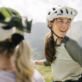Eine lächelnde Frau mit Fahrradhelm genießt die Berglandschaft bei den 360 Flow Trails in Kitzbühel, während sie sich mit Freunden unterhält.