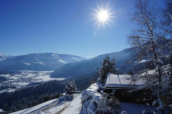 Verschneite Berghütte vor einem weiten Tal mit Bergen unter blauem Himmel und strahlender Sonne.