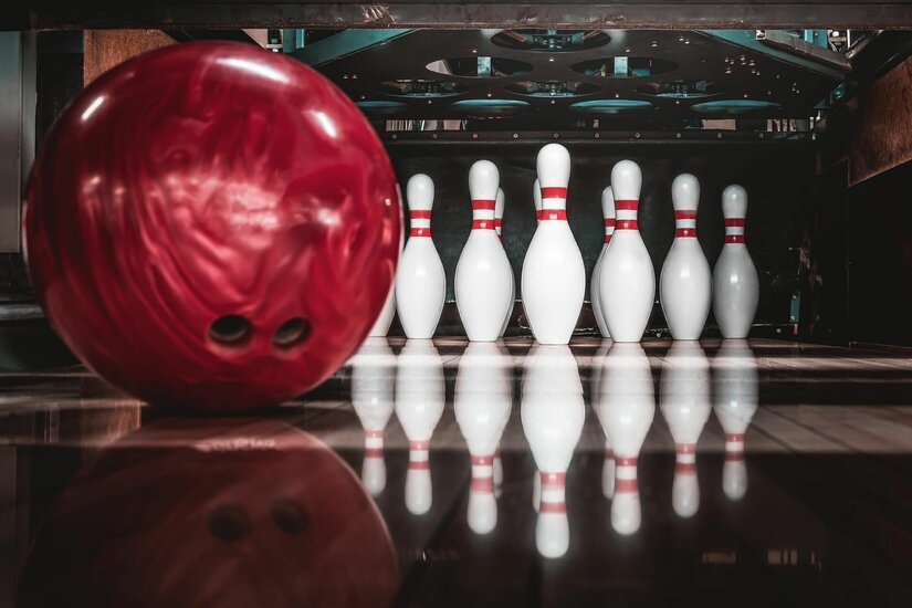 Rote Bowlingkugel im Vordergrund vor aufgestellten weißen Pins auf einer Bowlingbahn.