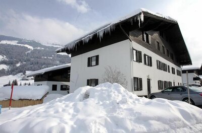 Mehrstöckiges weißes Gebäude mit dunklem Holzdach und Eiszapfen vor verschneiten Bergen.