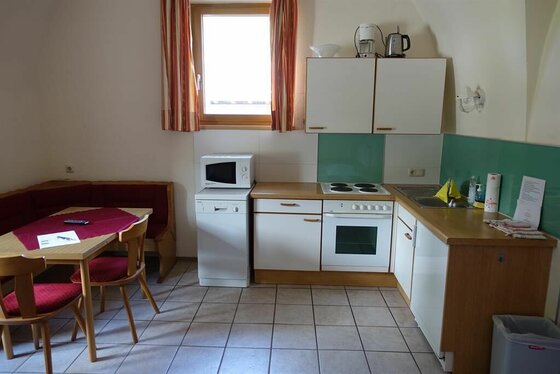 Kleine Einbauküche mit weißer Front, Elektroherd und angrenzender Sitzecke mit Holztisch.