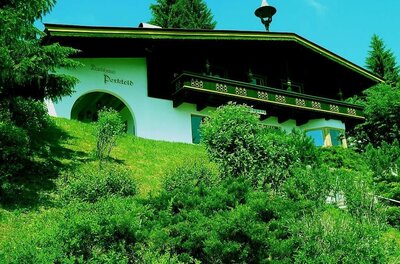 Zweistöckiges Landhaus mit dunklem Holzbalkon auf einem grünen Hügel vor Nadelbäumen.