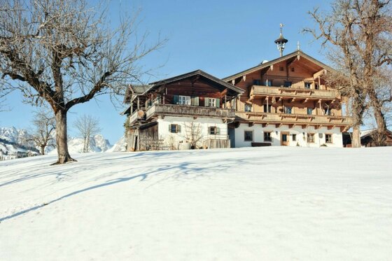 Traditioneller Bauernhof aus Holz und Stein in einer verschneiten Winterlandschaft unter blauem Himmel.
