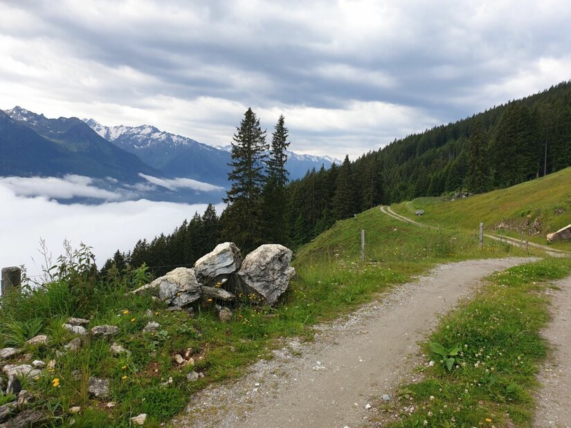 Schotterweg am Berghang mit Blick auf ein Wolkenmeer im Tal und schneebedeckte Gipfel.