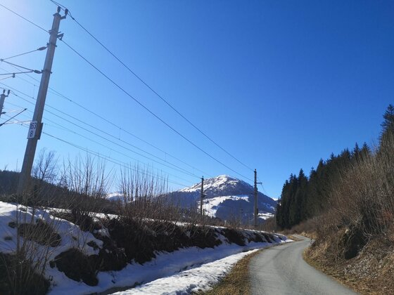 Wegverlauf an der Bahnlinie Asphaltierter Weg neben einer Bahnlinie mit Blick auf einen schneebedeckten Berg unter blauem Himmel.
