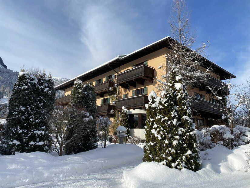 Das EDELWEISS KITZBÜHEL im Winter Mehrstöckiges Hotelgebäude mit Holzbalkonen in einer verschneiten Winterlandschaft unter blauem Himmel.