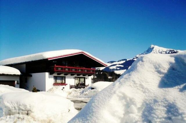 Winter Ein gemütliches Haus in einer verschneiten Landschaft unter klarem blauen Himmel. Hohe Schneehügel umgeben das Gebäude, während Berge im Hintergrund sichtbar sind.