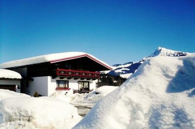 Ein gemütliches Haus in einer verschneiten Landschaft unter klarem blauen Himmel. Hohe Schneehügel umgeben das Gebäude, während Berge im Hintergrund sichtbar sind.