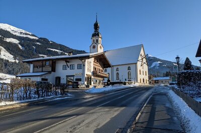 Verschneite Dorfstraße in Jochberg mit Blick auf die Pfarrkirche und traditionelle Alpenhäuser vor Bergkulisse.