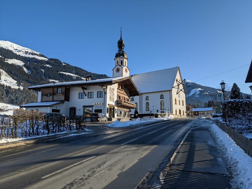 Ortszentrum Jochberg im Winter Verschneite Dorfstraße in Jochberg mit Blick auf die Pfarrkirche und traditionelle Alpenhäuser vor Bergkulisse.