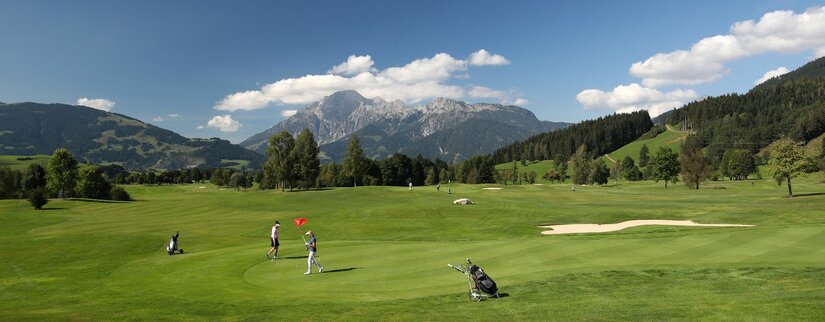 Golf Club Urslautal Golfplatz mit Spielern auf dem Green vor einer alpinen Bergkulisse unter blauem Himmel.