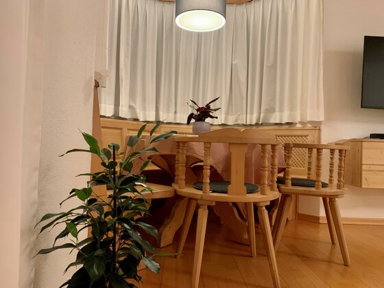 Rustikale Sitzecke aus Holz mit rundem Tisch, Stühlen und einer Eckbank vor einem Fenster mit weißen Vorhängen.