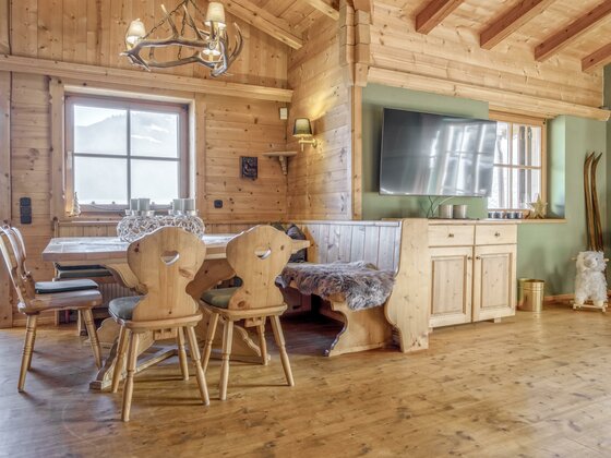 Rustikaler Essbereich aus hellem Holz mit Eckbank, Stühlen, Geweihlampe und einem Fernseher an der Wand.