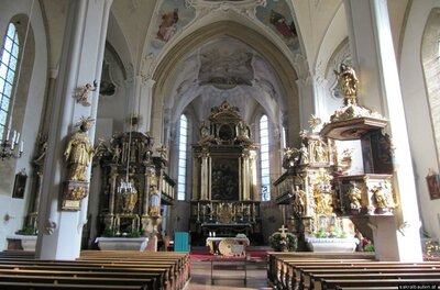 Ein schöner Kircheninnenraum mit kunstvollen Altären und einer hohen Decke. Die Bänke sind aufgereiht und der Altarbereich ist zentral sichtbar.